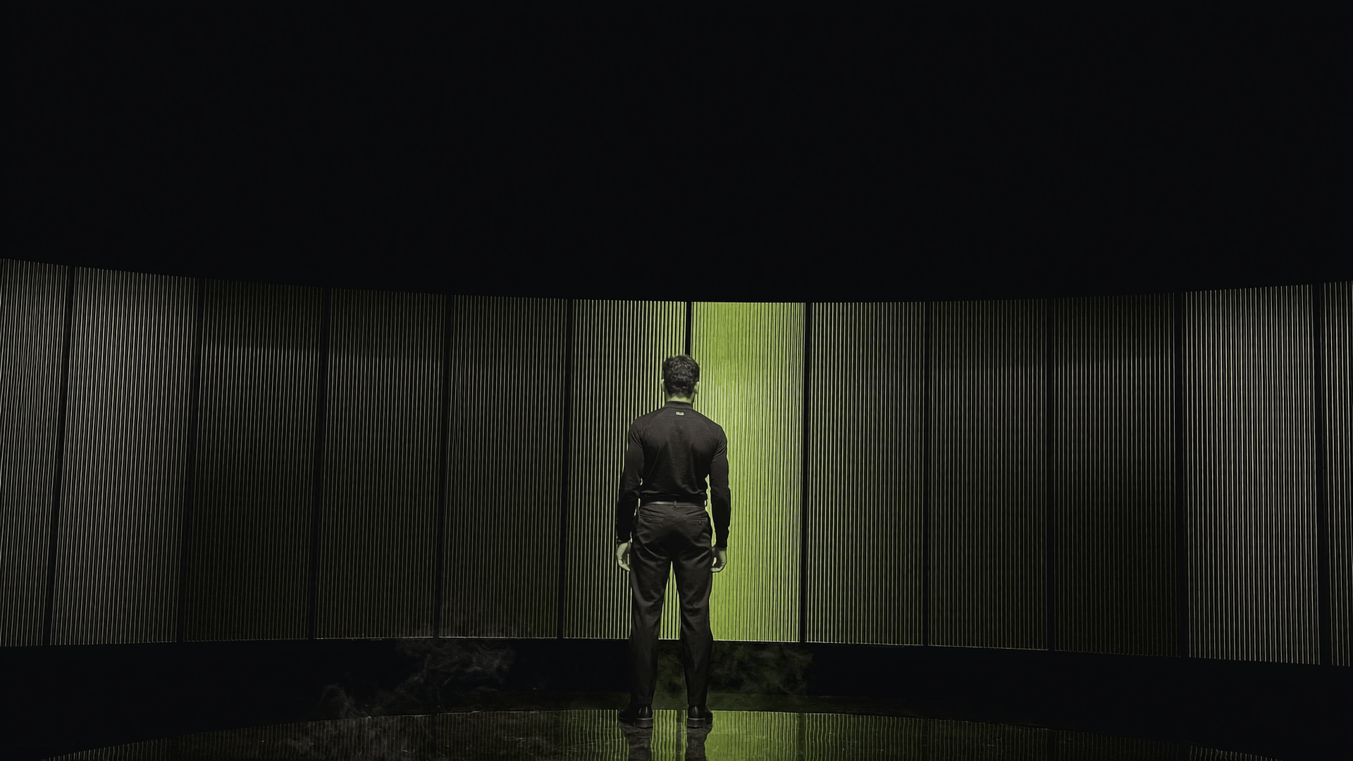 Kassem Kazal stands facing a wall of ribbed glass lit by a single olive-green panel