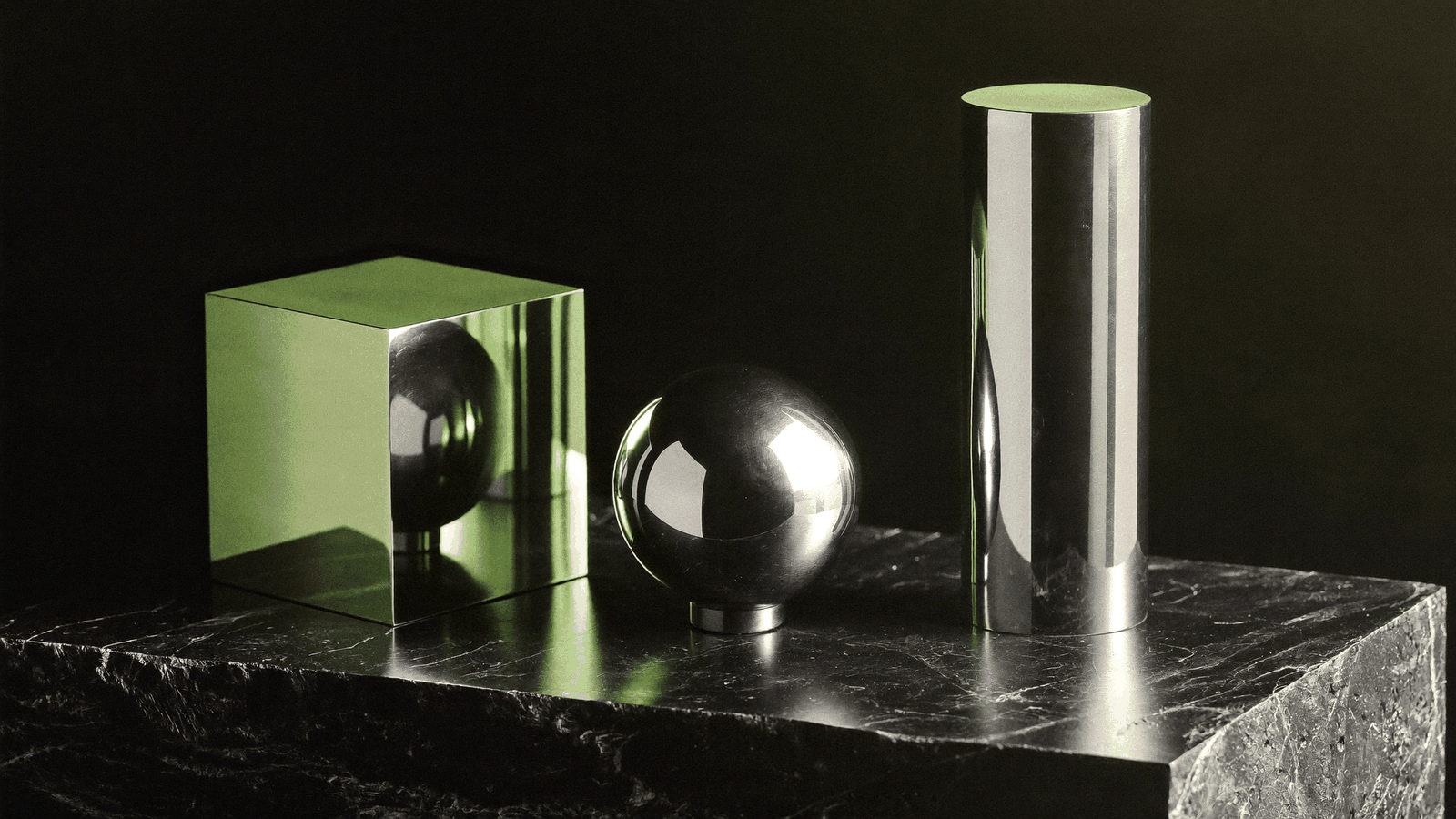 A chrome cube, sphere and cylinder lined up on dark stone