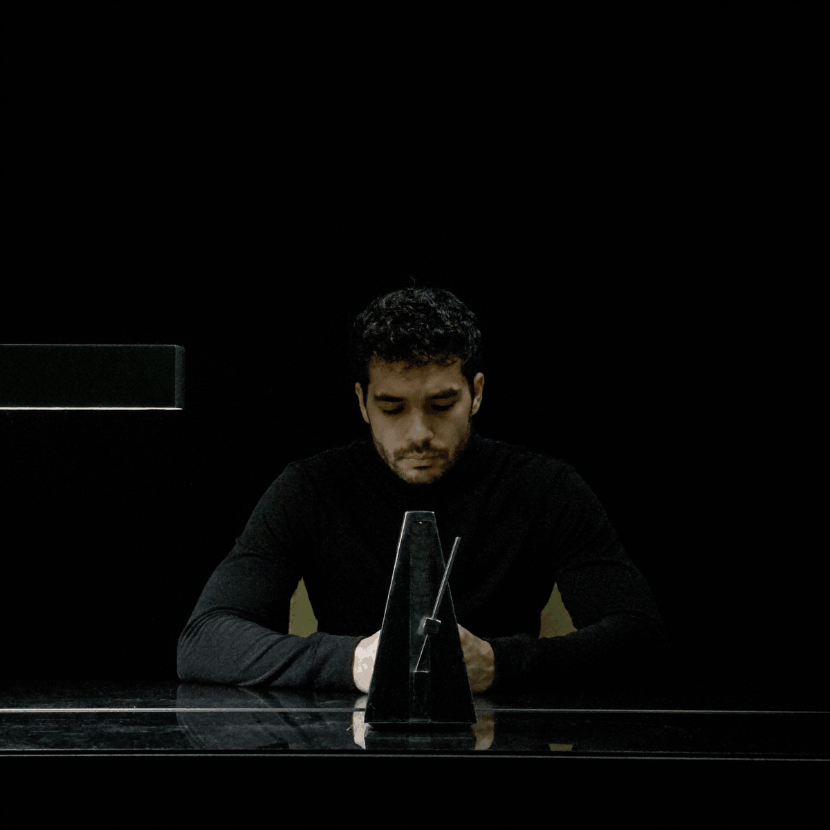 Kassem Kazal sits at a dark desk facing a metronome frozen mid-swing